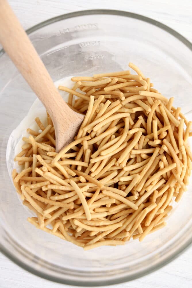 Overhead view of chow mein noodles being mixed into melted white chocolate in clear bowl with wooden spoon