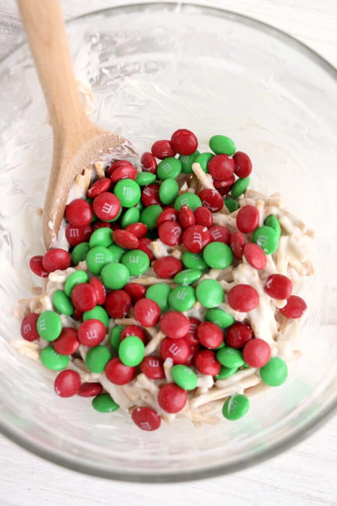 Overhead view of green and red M&Ms being stirred into ingredients in clear bowl with wooden spoon