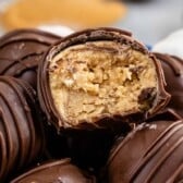 Close-up of chocolate truffles, one with a bite taken out showing a creamy peanut butter filling. The truffles, inspired by a classic peanut butter balls recipe, are drizzled with chocolate and stacked together.