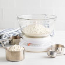 Flour in a glass mixing bowl on top of a kitchen scale with measuring cups off to the side