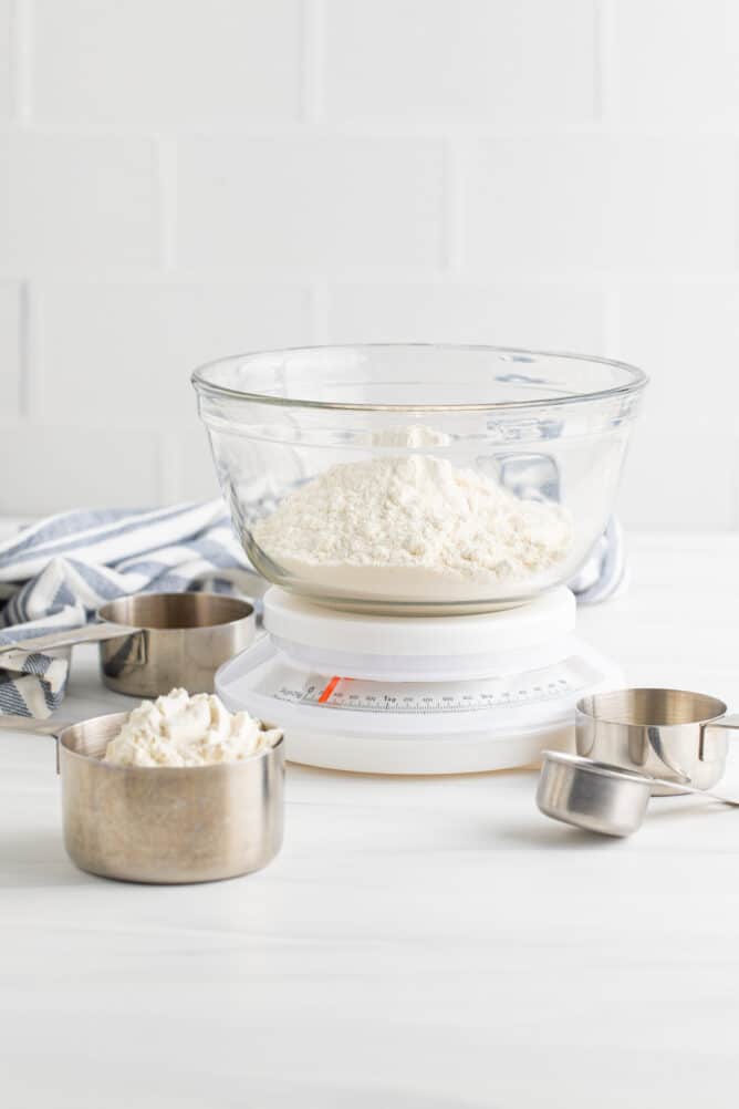 Flour in a glass mixing bowl on top of a kitchen scale with measuring cups off to the side