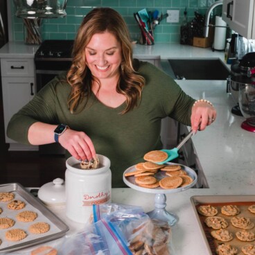 woman in green shirt standing at kitchen counter placing cookie in cookie jar with cookies all around on counter