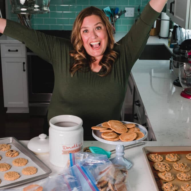 woman in green shirt standing at kitchen counter jumping for joy with cookies all around on counter