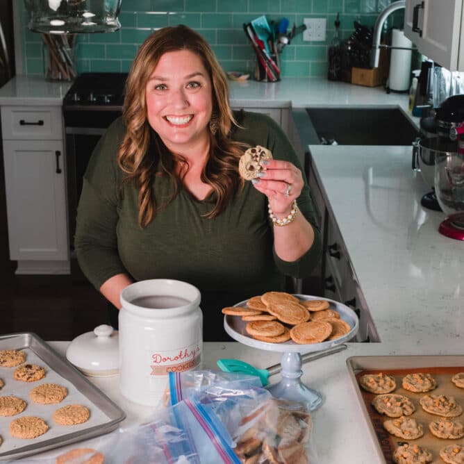 woman in green shirt standing at kitchen counter placing cookie in cookie jar with cookies all around on counter