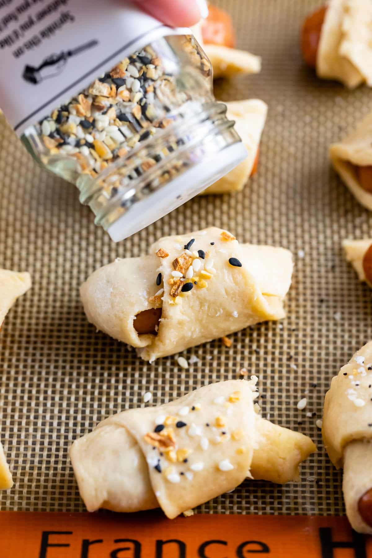 raw pig in a blanket on cookie sheet with bagel seasoning sprinkling on top.