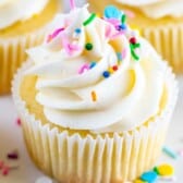 A Vanilla Cupcake with white frosting, decorated with colorful sprinkles, sits in a white paper liner. Other cupcakes and sprinkles are visible in the background, creating a delightful and sweet display.