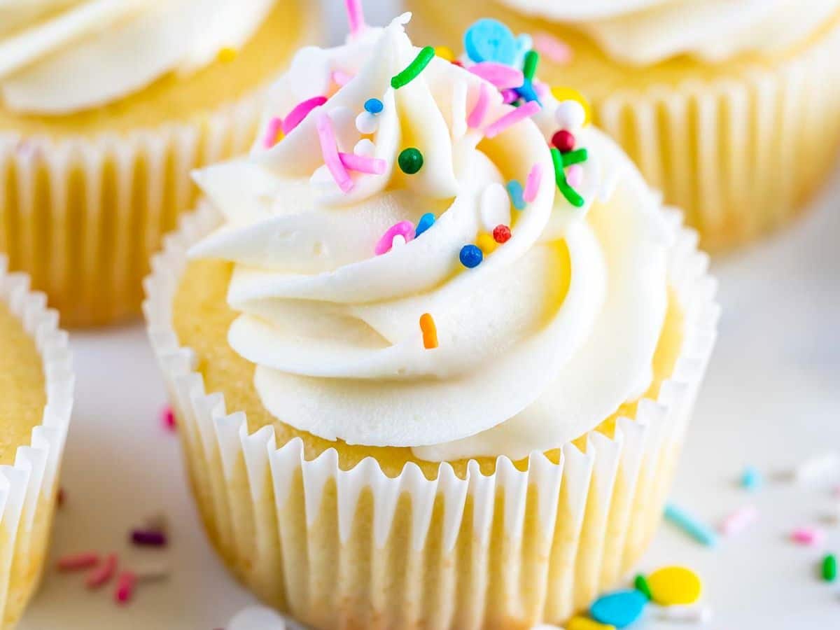 A Vanilla Cupcake with white frosting, decorated with colorful sprinkles, sits in a white paper liner. Other cupcakes and sprinkles are visible in the background, creating a delightful and sweet display.