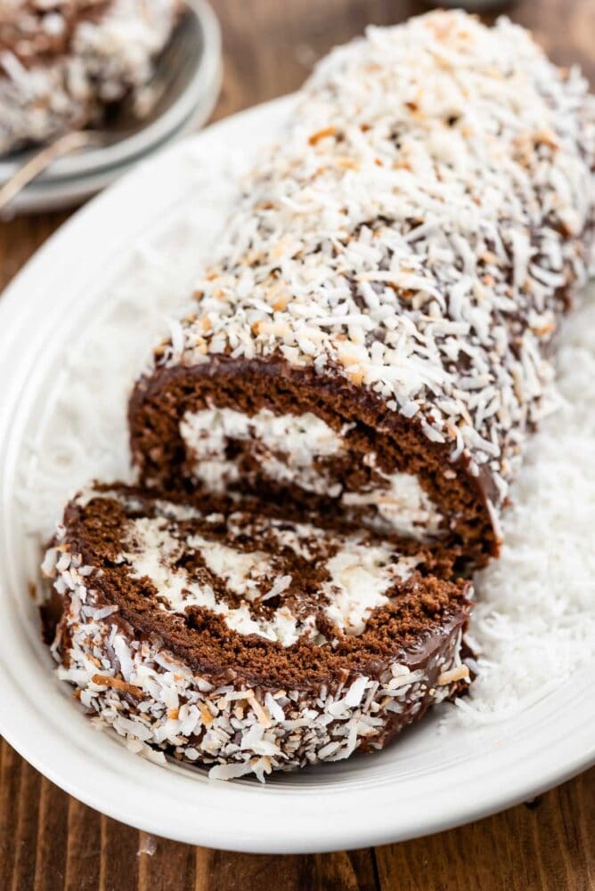 Chocolate coconut cake roll on a plate with one slice cut off and shredded coconut all around