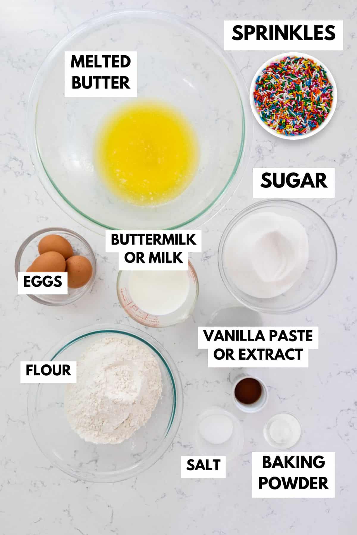 A collection of funfetti cupcake ingredients is arranged on a marble surface. Items include melted butter, sprinkles, eggs, flour, sugar, buttermilk or milk, vanilla paste or extract, salt, and baking powder, each labeled.