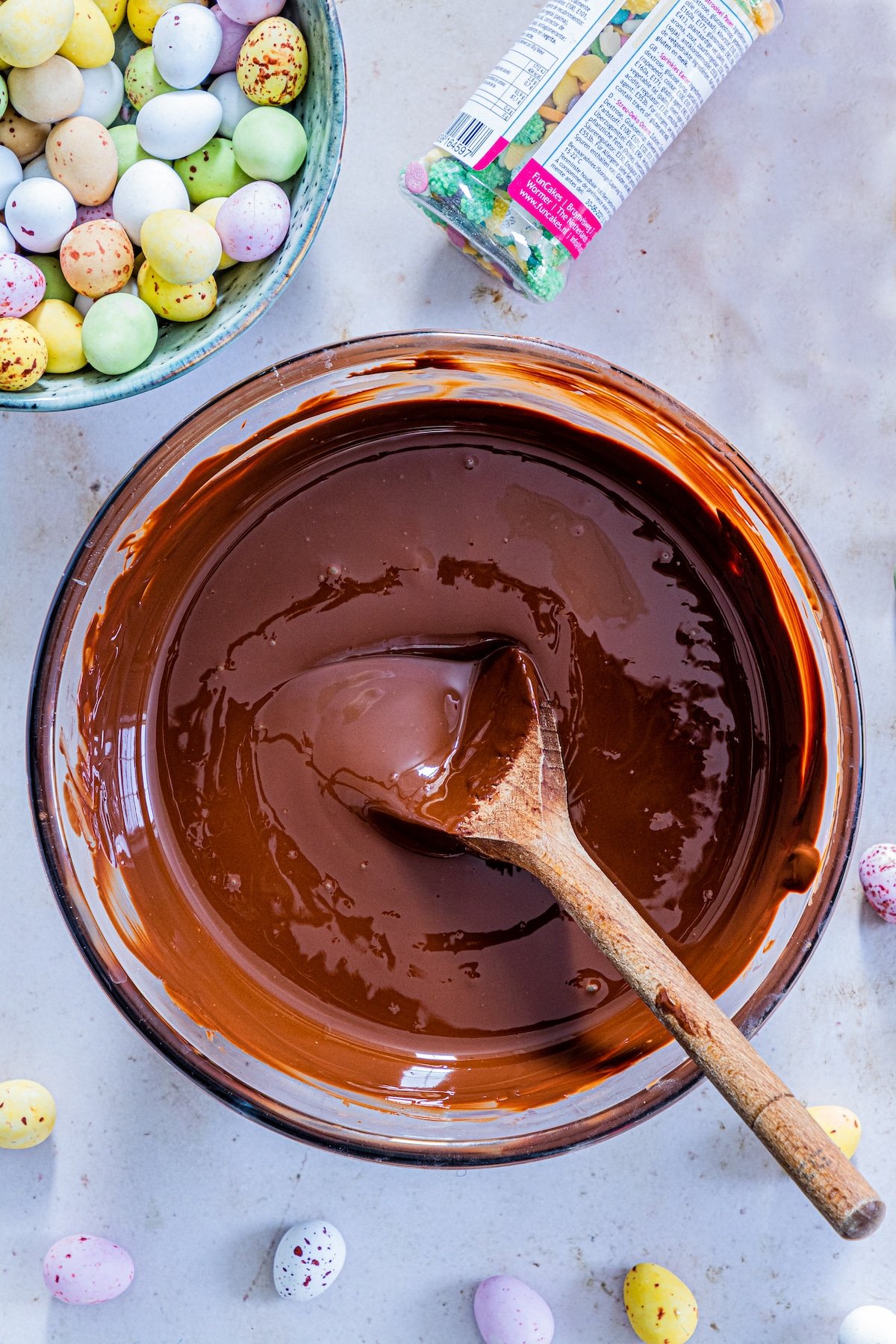 Overhead view of melted chocolate in a large bowl next to sprinkles and candy eggs