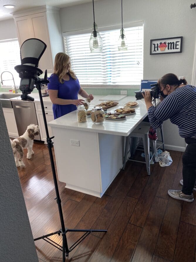 photographer taking picture of woman in kitchen with cookies all over white counter
