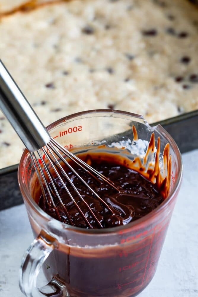 Close up shot of chocolate ganache in a measuring cup with chocolate chip banana bars in background