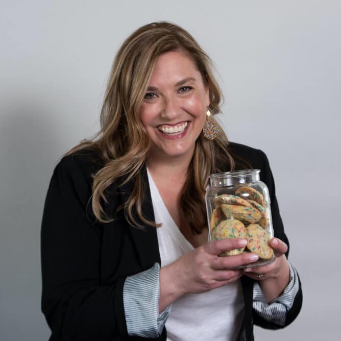 woman smiling wearing blazer and holding cookie jar full of sprinkle cookies