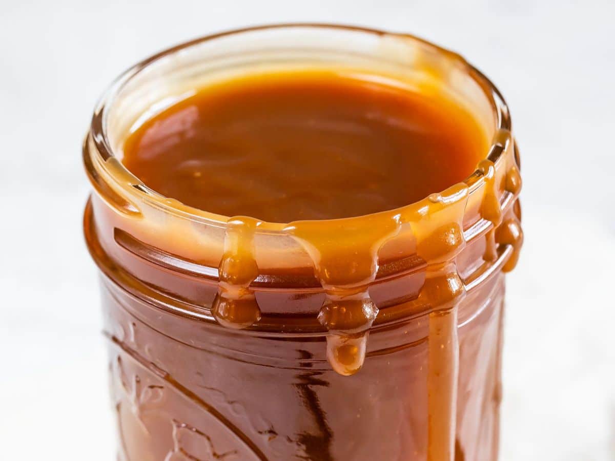 An open glass jar filled with thick caramel sauce, made from a classic caramel recipe, with caramel dripping down the sides.