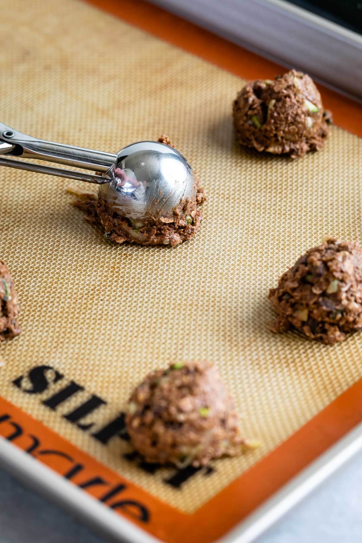 Scoops of chocolate zucchini cookies being placed on a baking sheet