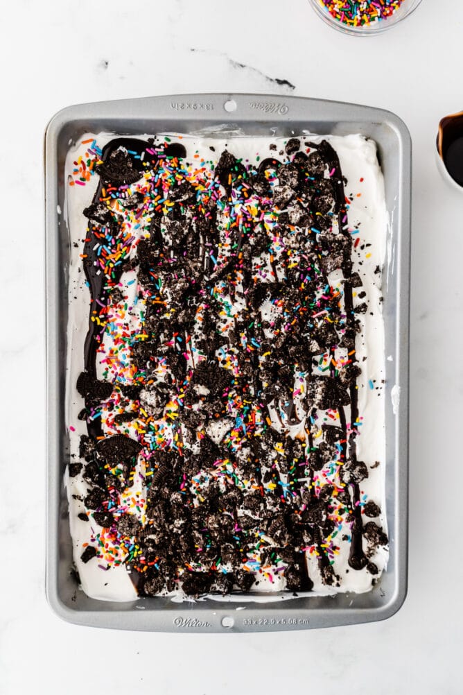 Overhead process shot showing ice cream sandwich cake topped with chocolate sauce and rainbow sprinkles