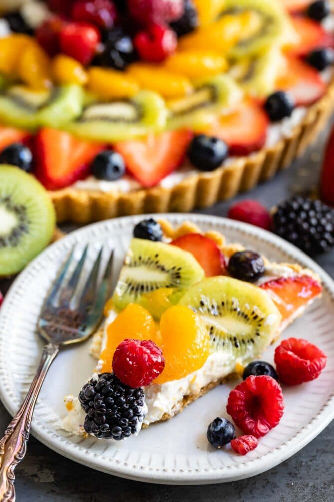 One slice of no bake fruit tart on a plate with more fruit and the rest of tart in the background