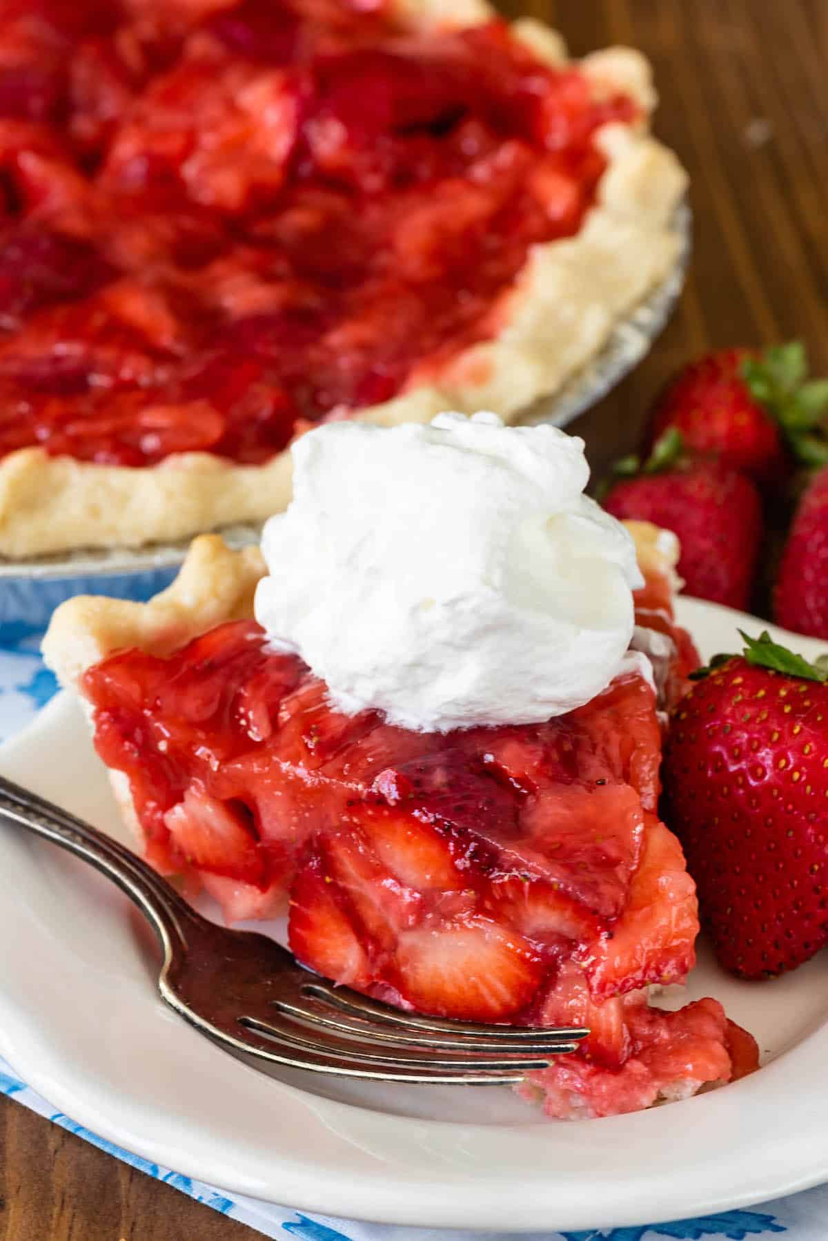 slice of strawberry pie on white plate with fork and dollup of cool whip with full pie behind.