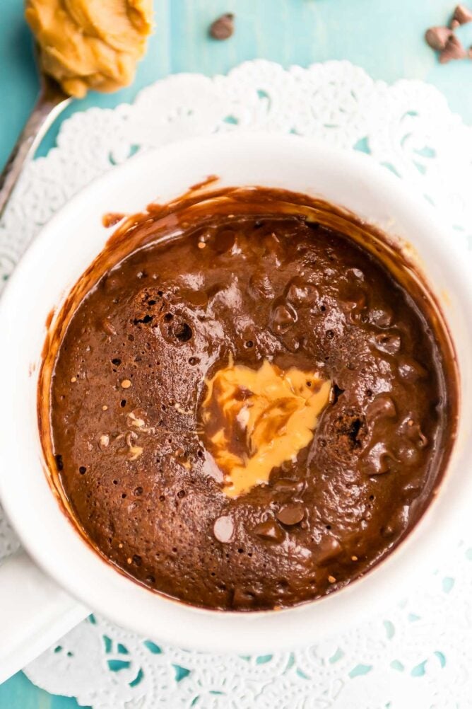 overhead shot of chocolate cookie in a white mug with peanut butter on top of a doilies and blue background