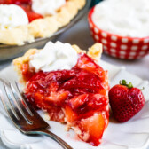 slice of strawberry pie on white plate with fork and dollup of cool whip with full pie behind.