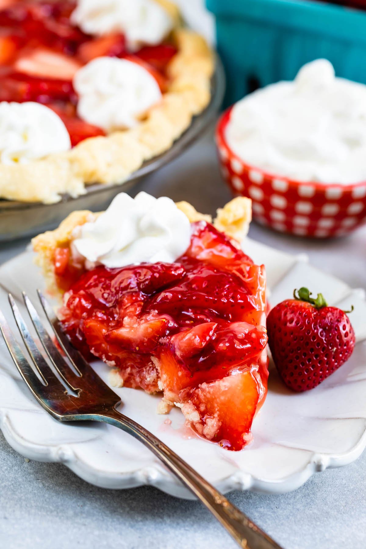 slice of strawberry pie on white plate with fork and dollup of cool whip with full pie behind.