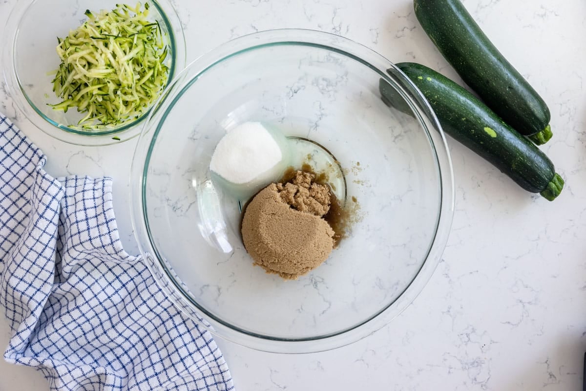 clear bowl with oil, brown sugar, and white sugar with zucchini around.