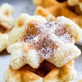 Close-up of two golden brown waffle cookies stacked on each other, dusted with powdered sugar. More waffles are visible blurred in the background.