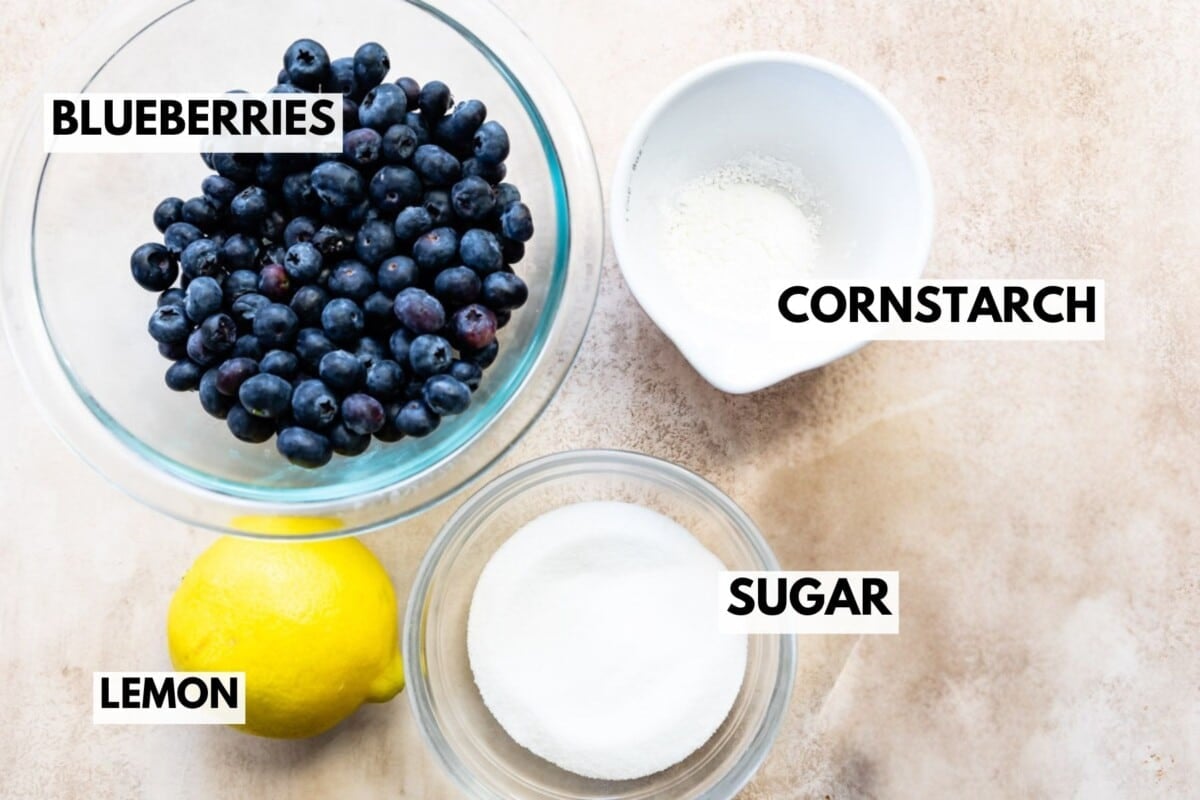 A bowl of blueberries, a lemon, a small bowl of sugar, and a small bowl of cornstarch—essential ingredients for Blueberry Crumble Pie—are labeled and arranged on a light countertop.