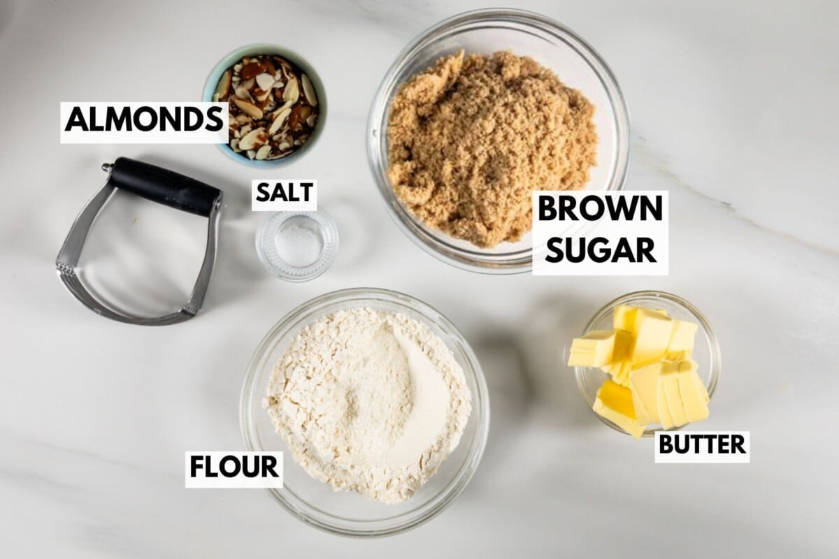Overhead view of labeled baking ingredients for Blueberry Crumble Pie on a white surface: a bowl of sliced almonds, brown sugar, small bowl of salt, flour, diced butter, and a pastry cutter.