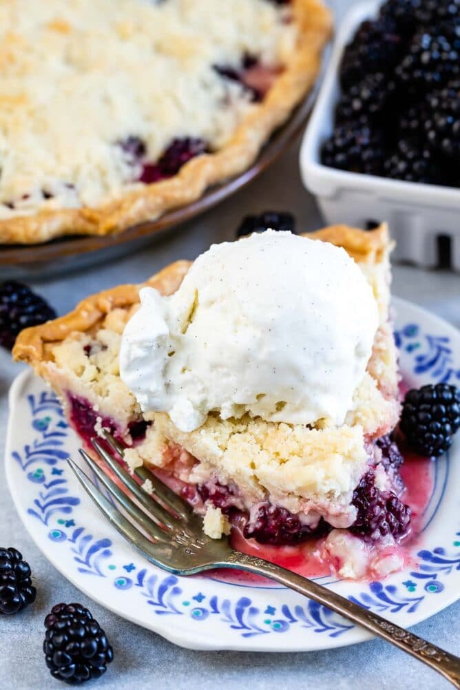 One slice of blackberry pie with crumble topping and a scoop of ice cream on top on a plate