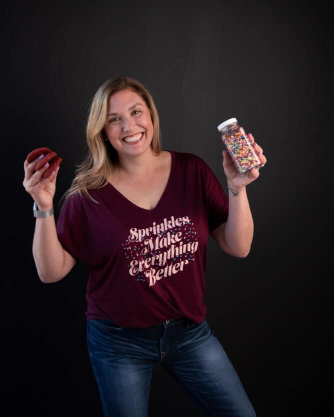 woman in maroon shirt holding sprinkles and cookies