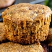 A close-up of a moist zucchini muffin with a golden-brown top, showing its fluffy, textured interior—perfectly highlighting why zucchini muffins are such a delicious, wholesome treat. More muffins are partially visible in the background.