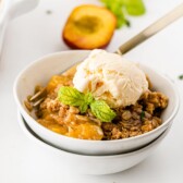 Peach crisp in a white bowl topped with vanilla ice cream and mint leaves