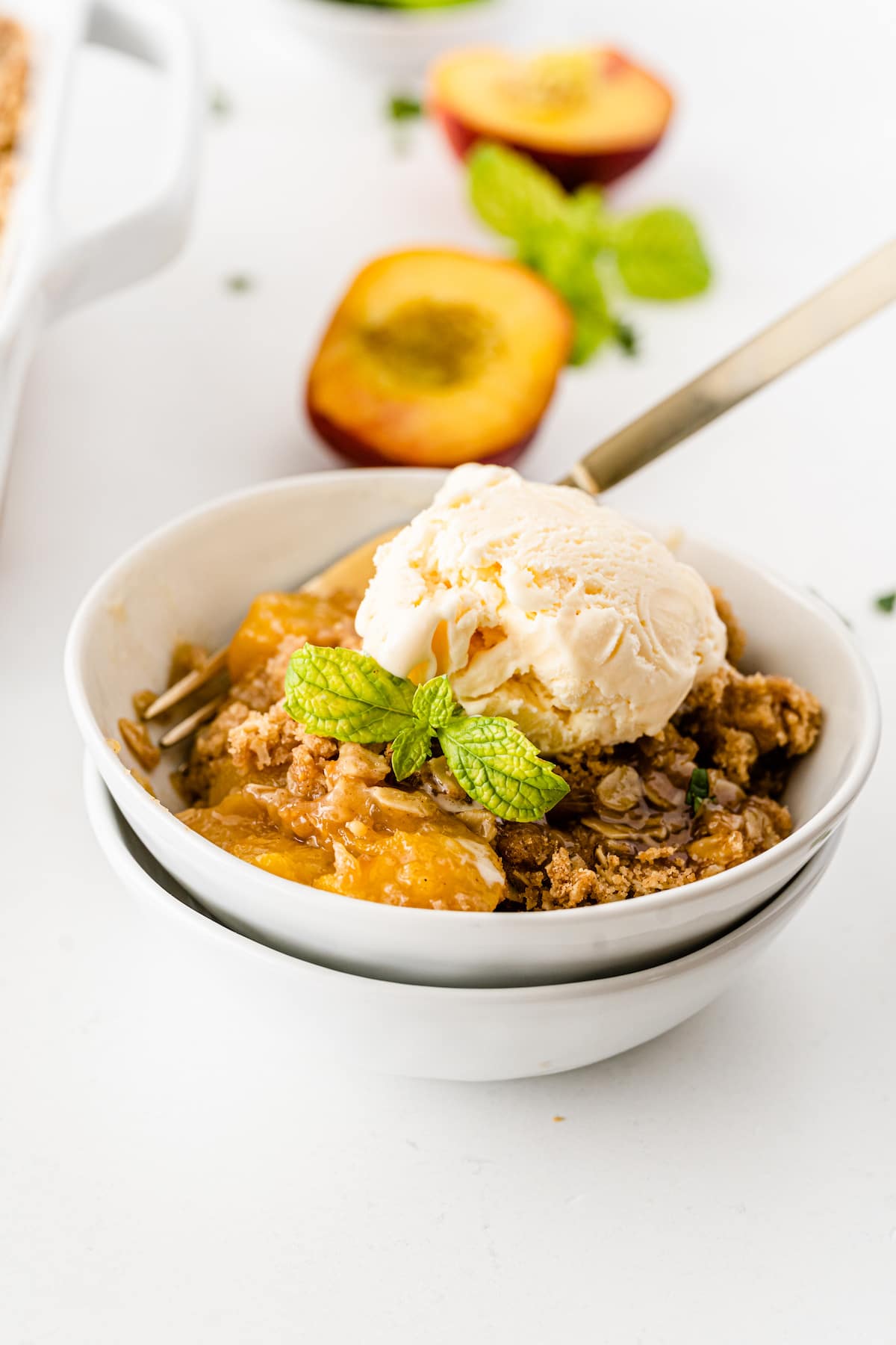 Peach crisp in a white bowl topped with vanilla ice cream and mint leaves