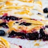 A close-up of a sliced blueberry danish with golden, flaky crust, filled with juicy blueberries and topped with a white icing drizzle. Fresh blueberries are scattered nearby on the white surface.