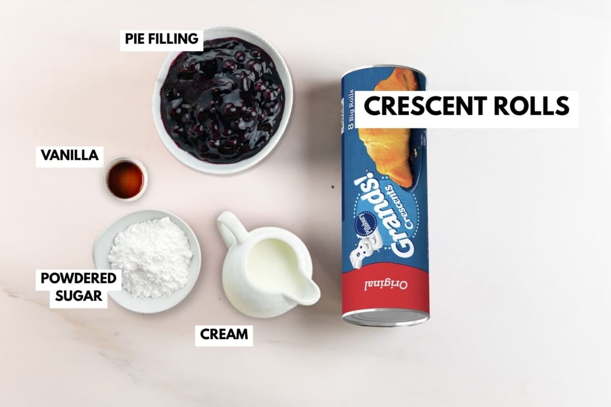 A can of Grands! crescent rolls, a bowl of pie filling for blueberry danish, a small bowl of vanilla, a dish of powdered sugar, and a small jug of cream are arranged on a light surface with labels for each ingredient.