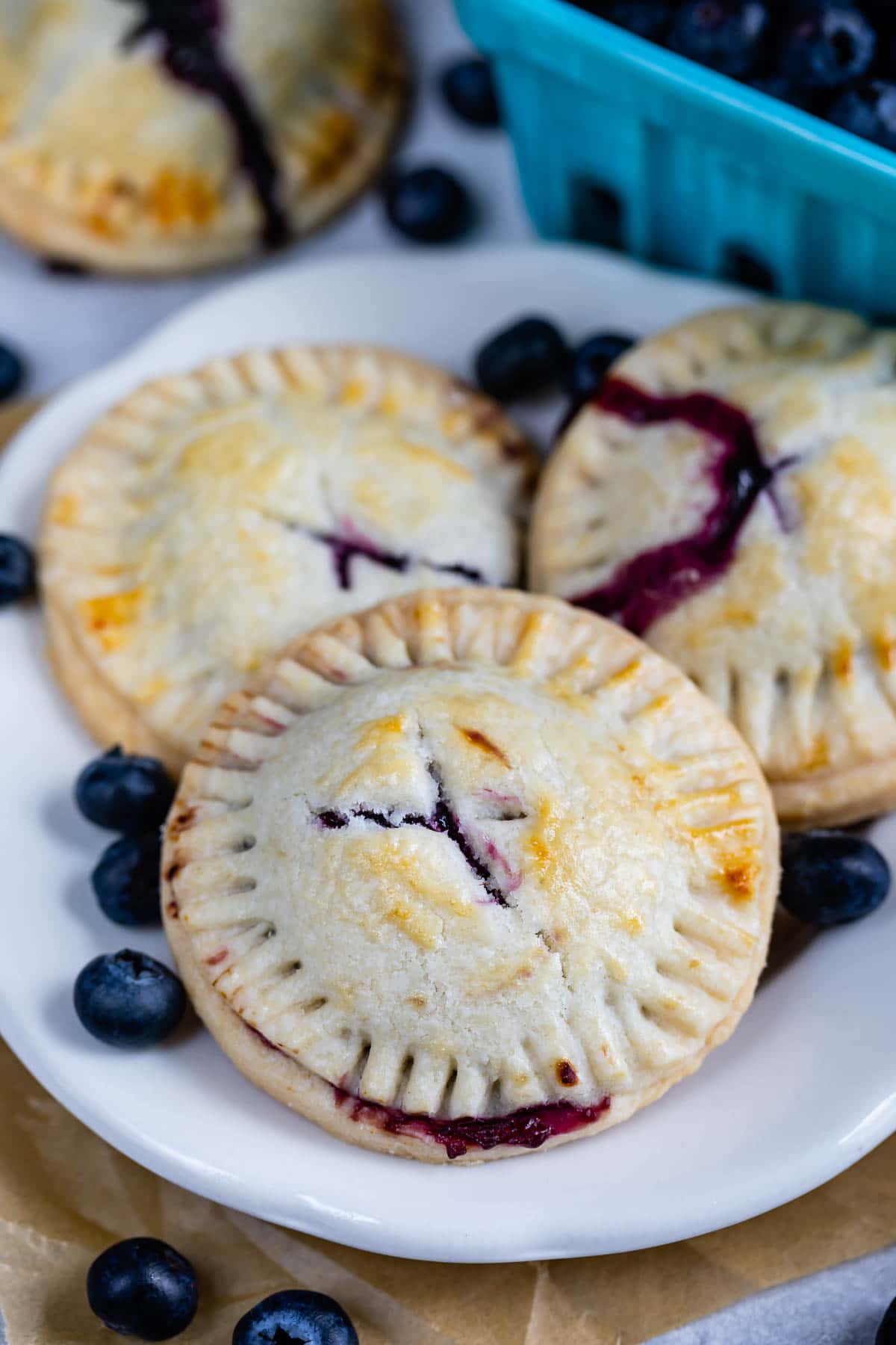 Three blueberry hand pies on a white plate with blueberries behind it