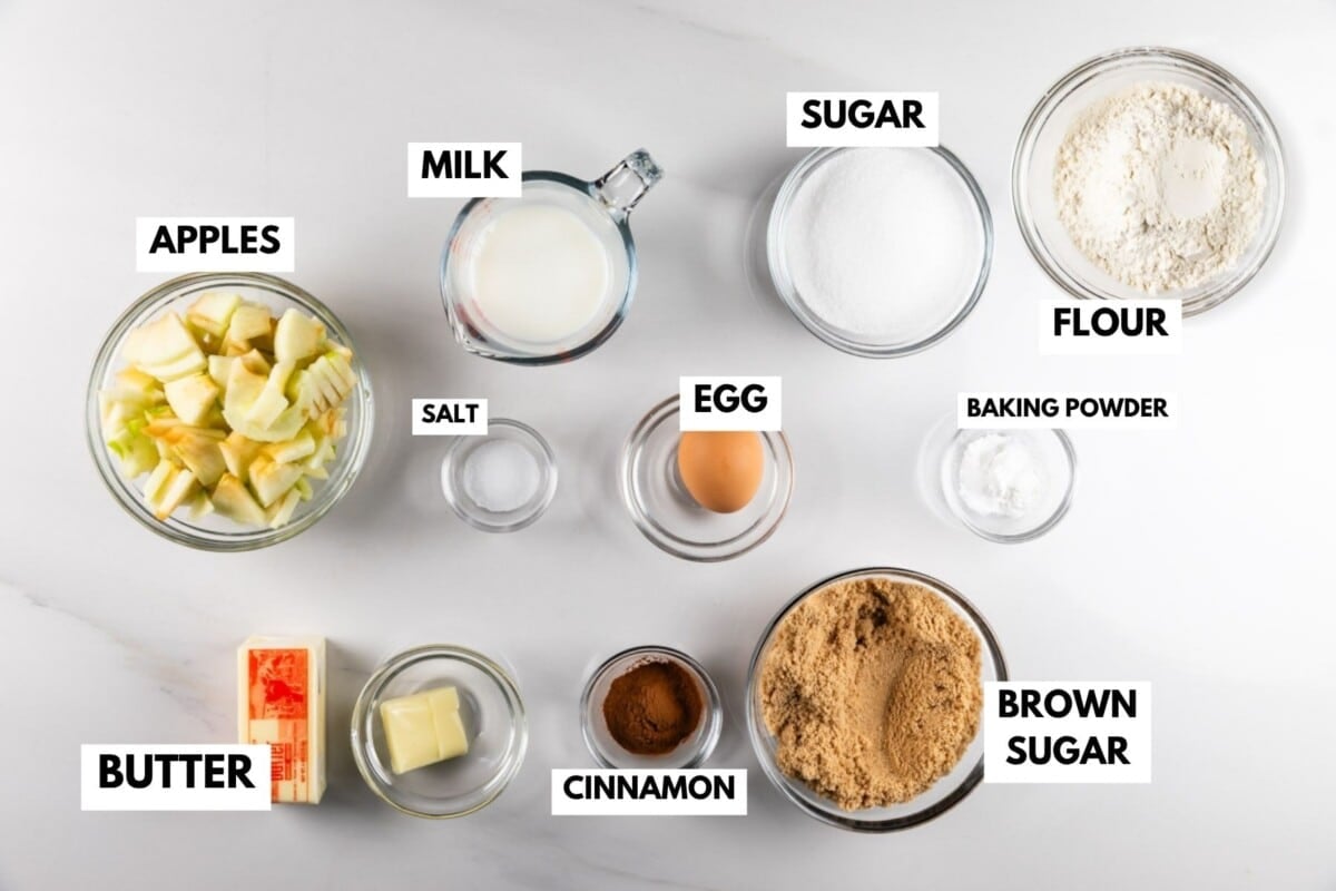 Labeled baking ingredients for apple coffee cake on a white surface: apples, butter, milk, salt, cinnamon, egg, sugar, flour, baking powder, and brown sugar in clear bowls and containers.