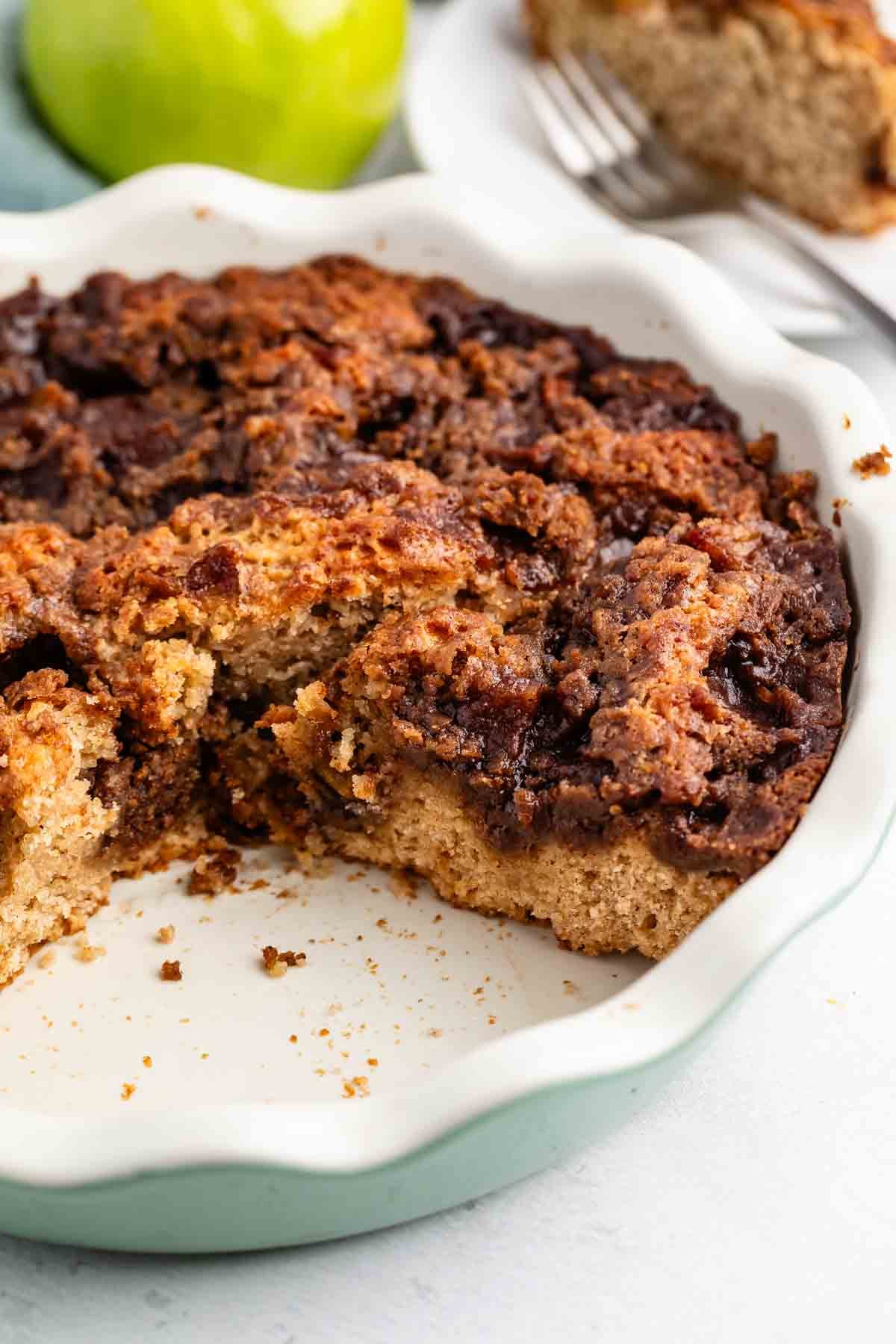 A close-up of a partially eaten apple coffee cake with a crumbly cinnamon topping in a white pie dish, with a green apple and a fork in the background.