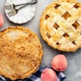 Two peach pies sit on a table next to whole peaches, a blue and white checkered cloth, and a white plate with two forks. One Peach Pie has a solid crust; the other features a classic lattice crust.