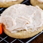 A frosted sugar cookie with creamy icing sits on a cooling rack. A small candy pumpkin is visible next to the cookie on a wooden surface, completing the festive look of these delightful Pumpkin Spice Cookies.
