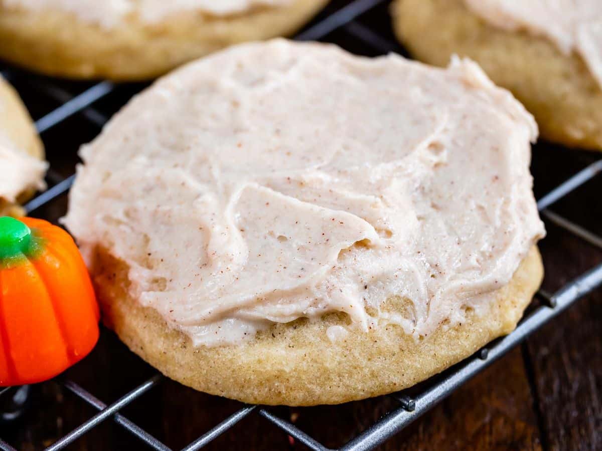 A frosted sugar cookie with creamy icing sits on a cooling rack. A small candy pumpkin is visible next to the cookie on a wooden surface, completing the festive look of these delightful Pumpkin Spice Cookies.