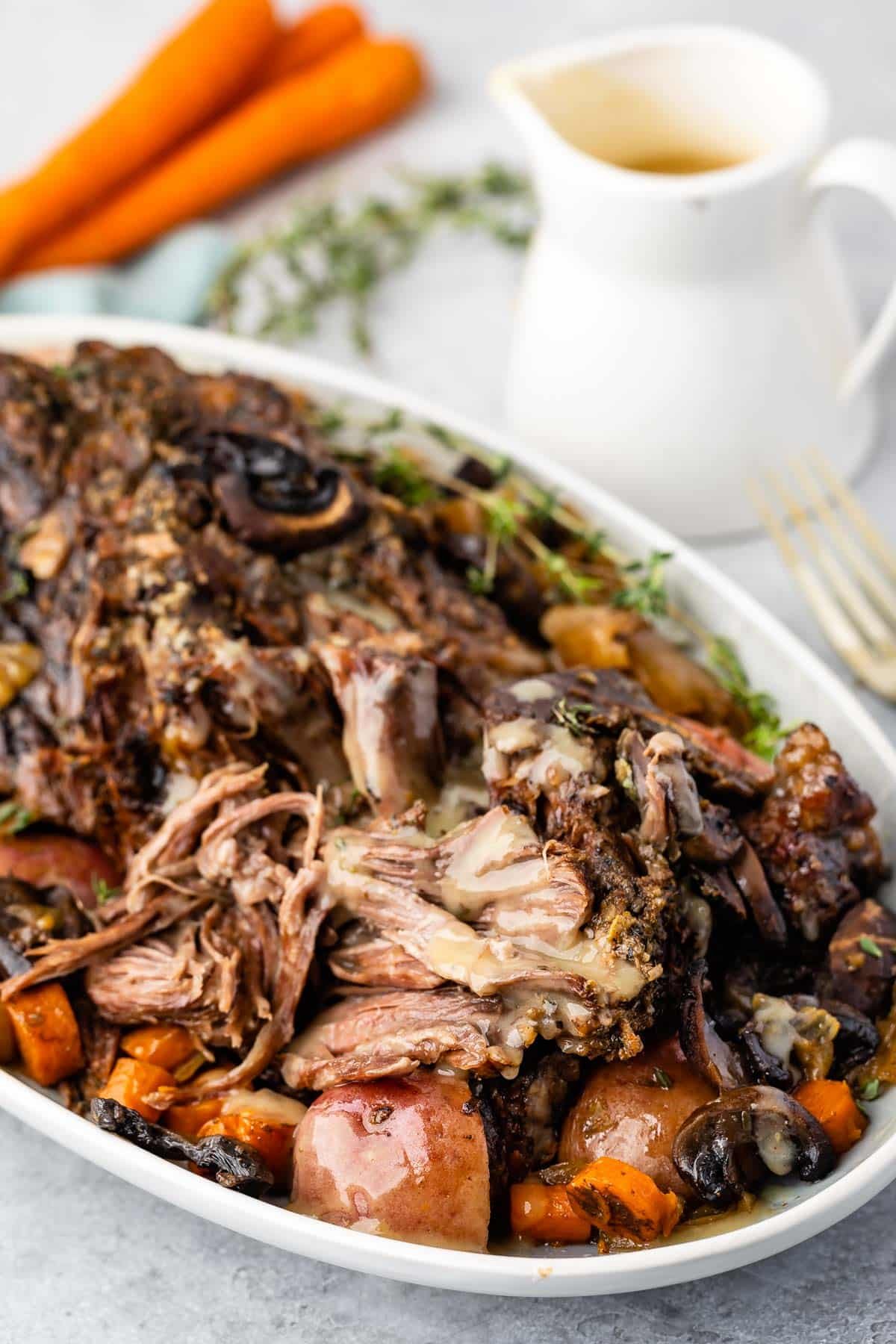 white serving platter with meat, potatoes and carrots around the edge with pitcher of gravy and carrots behind