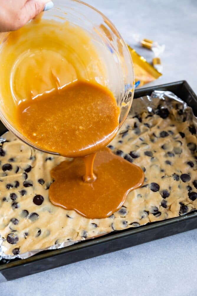Caramel filling being poured over chocolate chip cookie layer