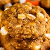 Pumpkin oatmeal cookies on a cutting board with pumpkin candies and white chocolate chips with recipe title on top of image