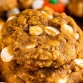 Close-up of soft, chewy pumpkin oatmeal cookies with visible white chocolate chips and oats, stacked on top of each other, with small pumpkin-shaped candies blurred in the background.