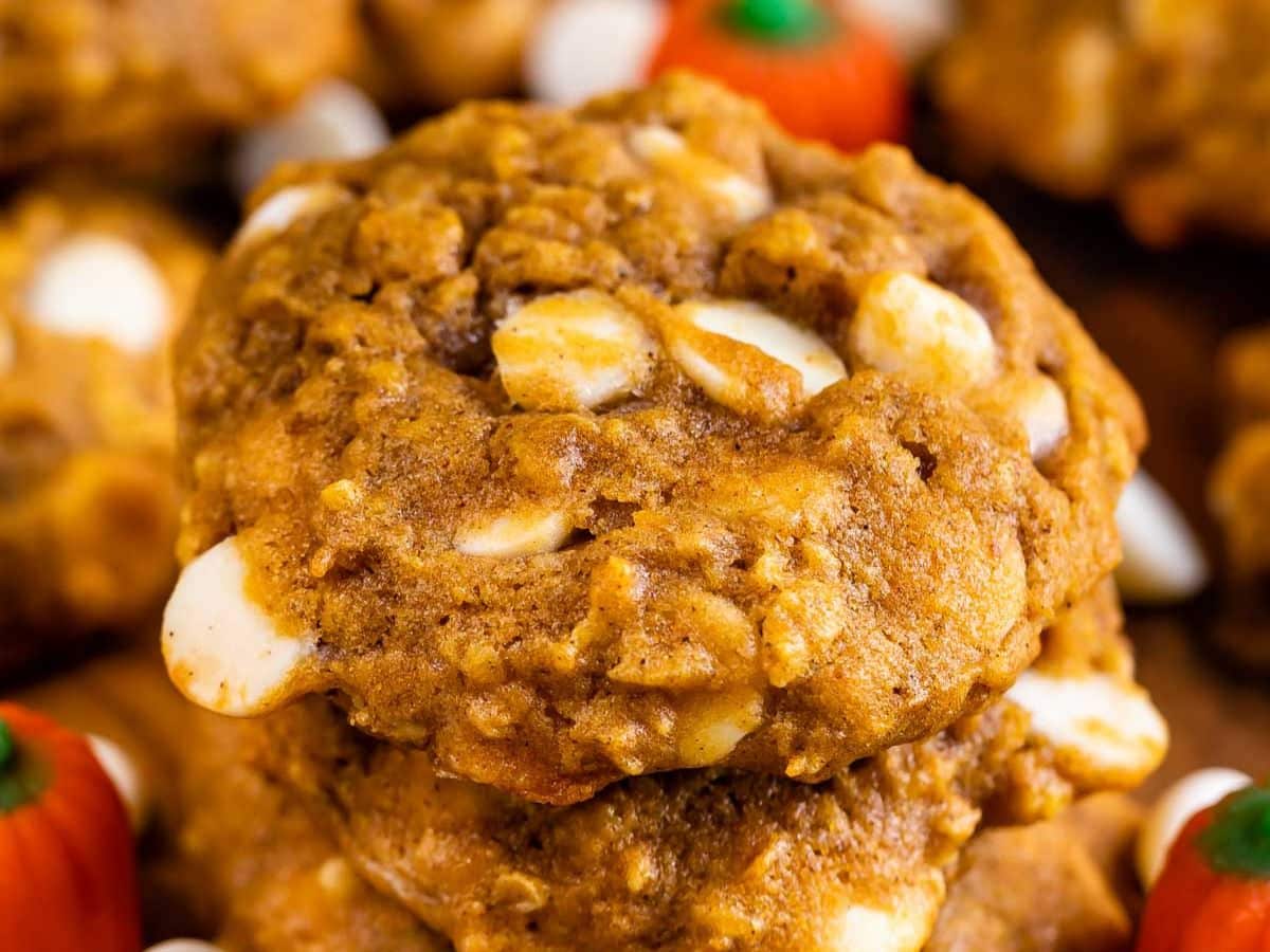 Close-up of soft, chewy pumpkin oatmeal cookies with visible white chocolate chips and oats, stacked on top of each other, with small pumpkin-shaped candies blurred in the background.