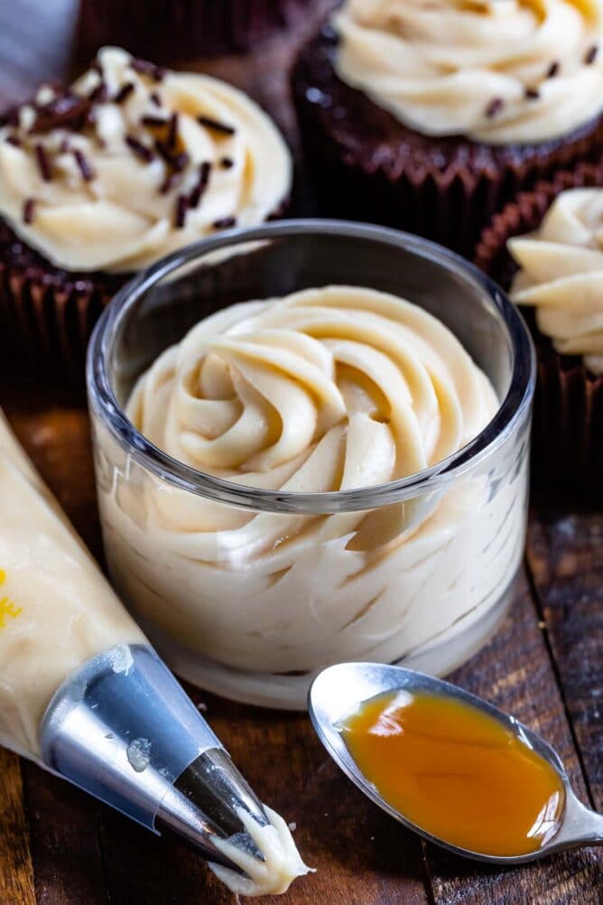 frosting in jar with caramel on spoon and piping bag in front and cupcakes behind