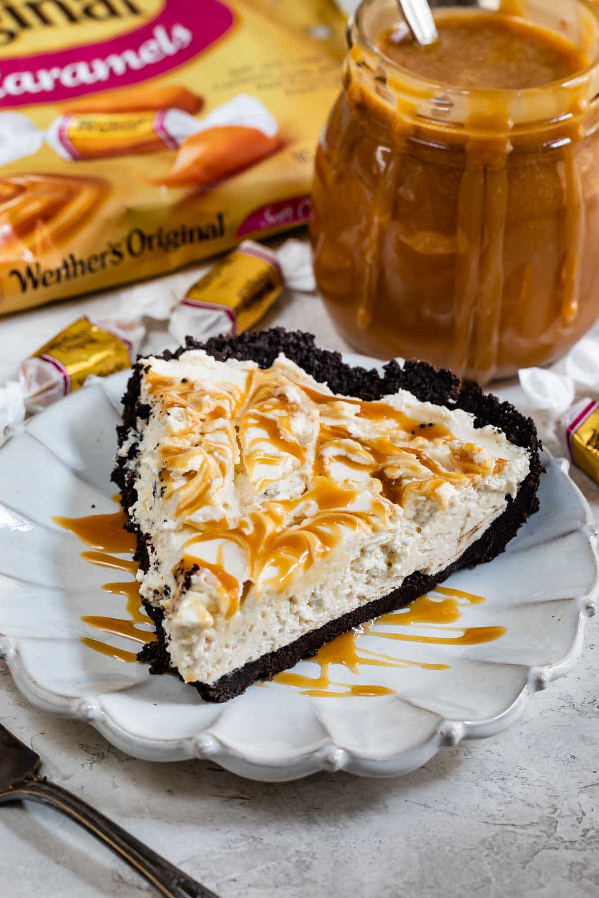 One slice of caramel cheesecake pie on a plate with caramel sauce and candies in the background
