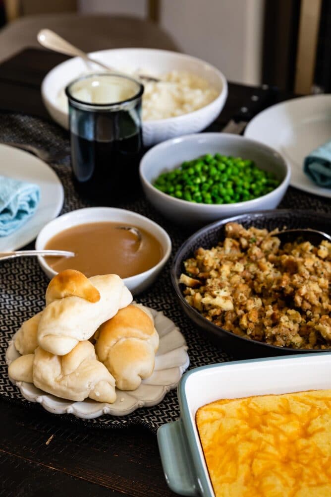 thanksgiving dinner on table - bowls of peas, stuffing, gravy, cornbread, rolls and mashed potatoes with place settings